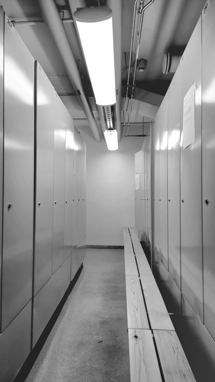 Black and white image of a locker room with a wooden bench and fluorescent lights.
