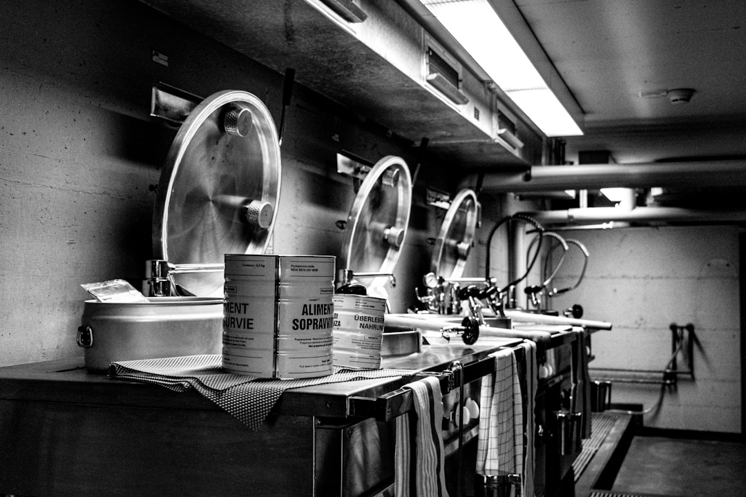Cold War kitchen inside the Sonnenberg bunker in Lucerne, Switzerland. Black and white photo of survival food cans, industrial cooking pots and stainless steel sinks used to prepare emergency rations in a civil defense shelter.